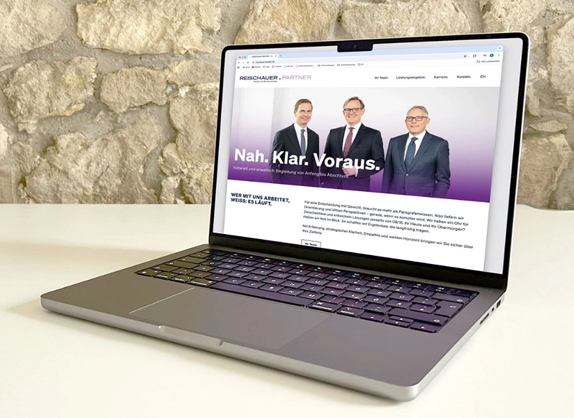 Die Startseite der Webseite Reischauer und Partner ist auf dem Bildschirm des MacBooks zu sehen. Das Laptop steht auf einen weißen Tische im Hintergrund ist eine unverputzte Wand zu sehen.