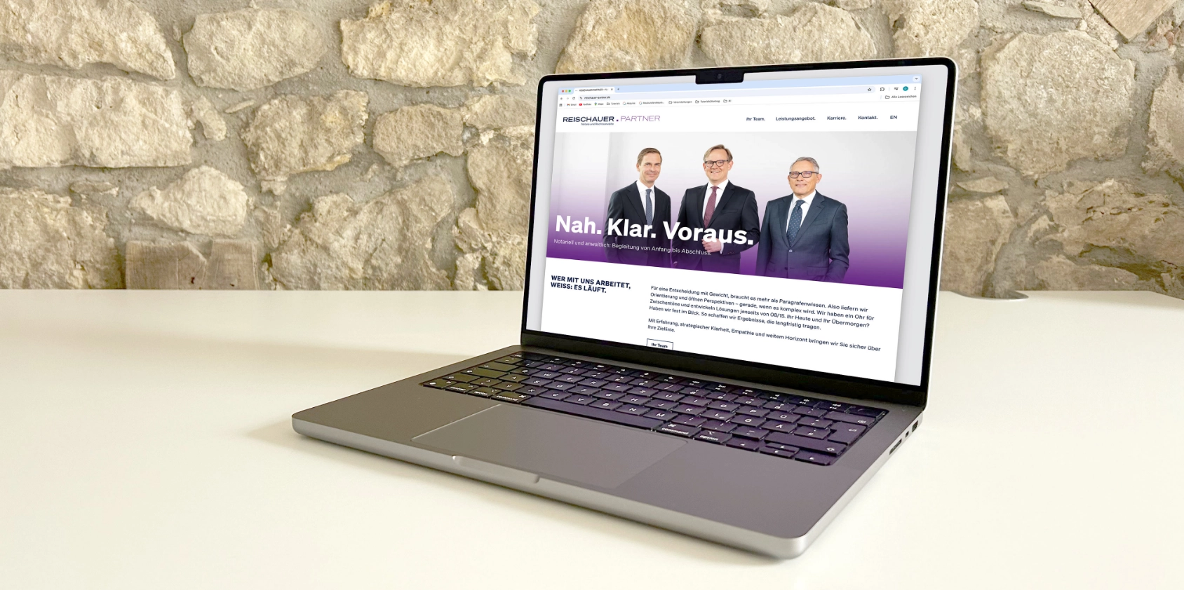 Die Startseite der Webseite Reischauer und Partner ist auf dem Bildschirm des MacBooks zu sehen. Das Laptop steht auf einen weißen Tische im HIntergrund ist eine unverputzte Wand zu sehen.